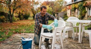 Homme nettoyant du mobilier de jardin en plastique blanc sur une terrasse avant l'hiver, dans un décor automnal.